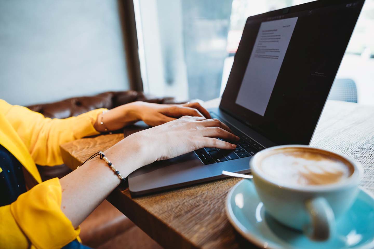 Hände auf einer Laptoptastatur, daneben eine Tasse Cappuccino
