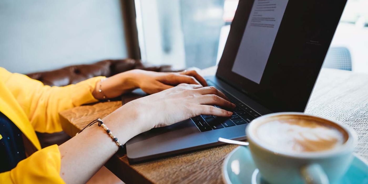 Hände auf einer Laptoptastatur, daneben eine Tasse Cappuccino